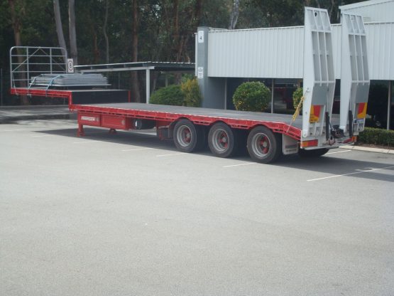 drop deck trailer not hooked onto truck in red standing in parking lot in front of gumtrees with loading tracks hoisted up
